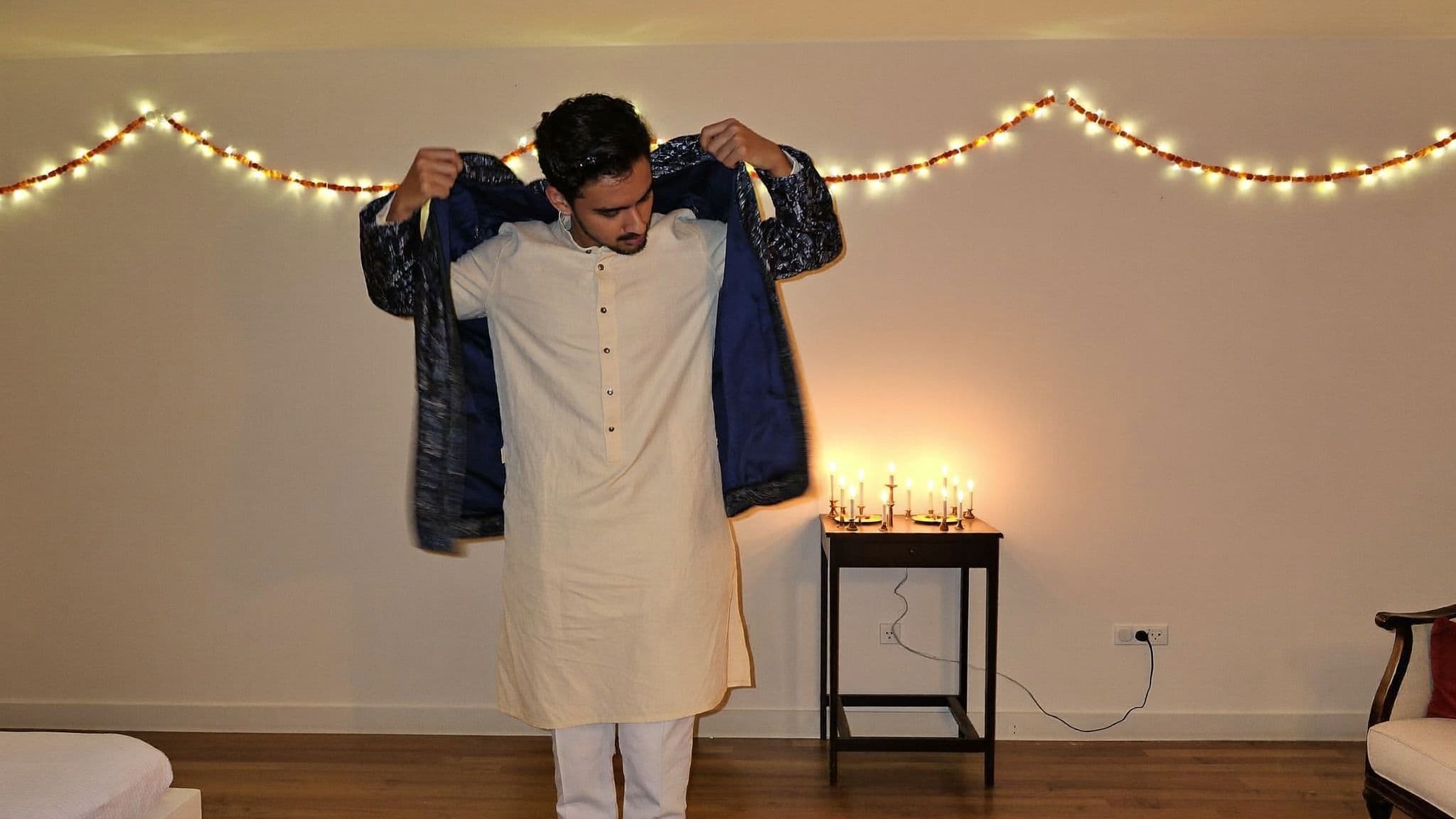 Man in an ivory kurta putting on a midnight-blue brocade Nehru jacket in a living room lit with string lights and candles