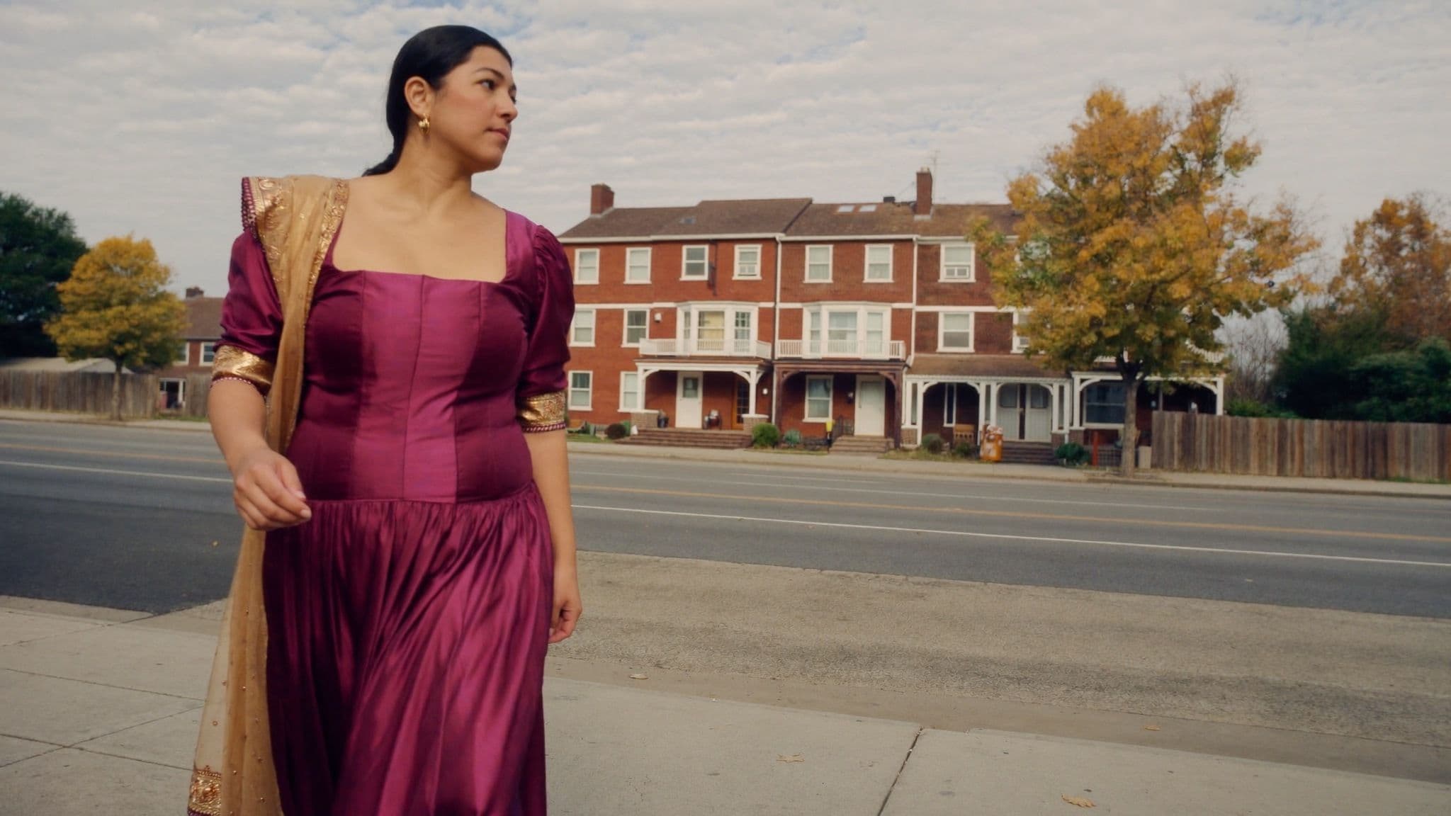 Woman in a plum silk fitted bodice and flowing skirt with a gold dupatta, walking past brick houses in autumn