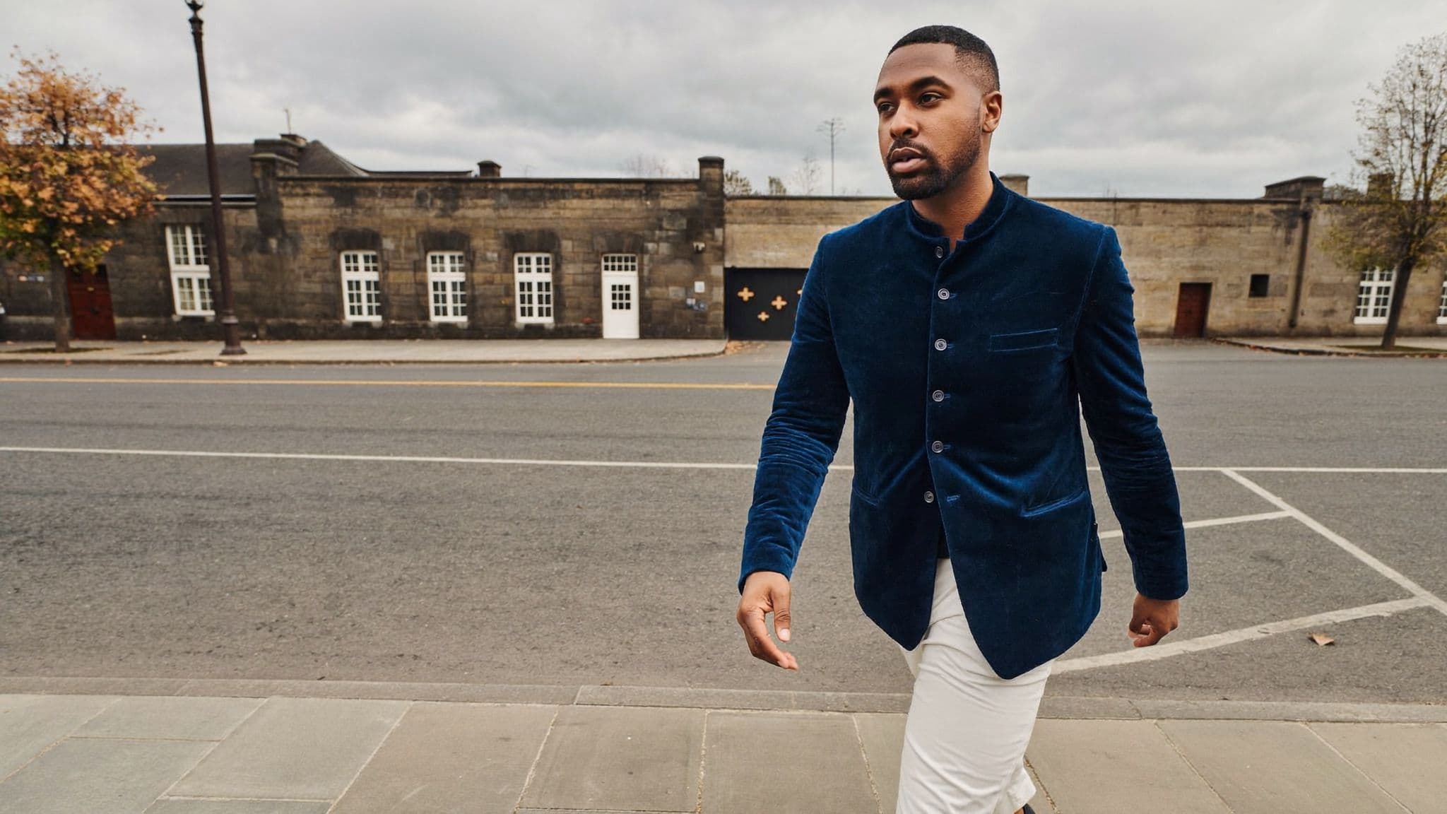 Man in a midnight-blue velvet bandhgala jacket and ivory trousers walking along a stone street
