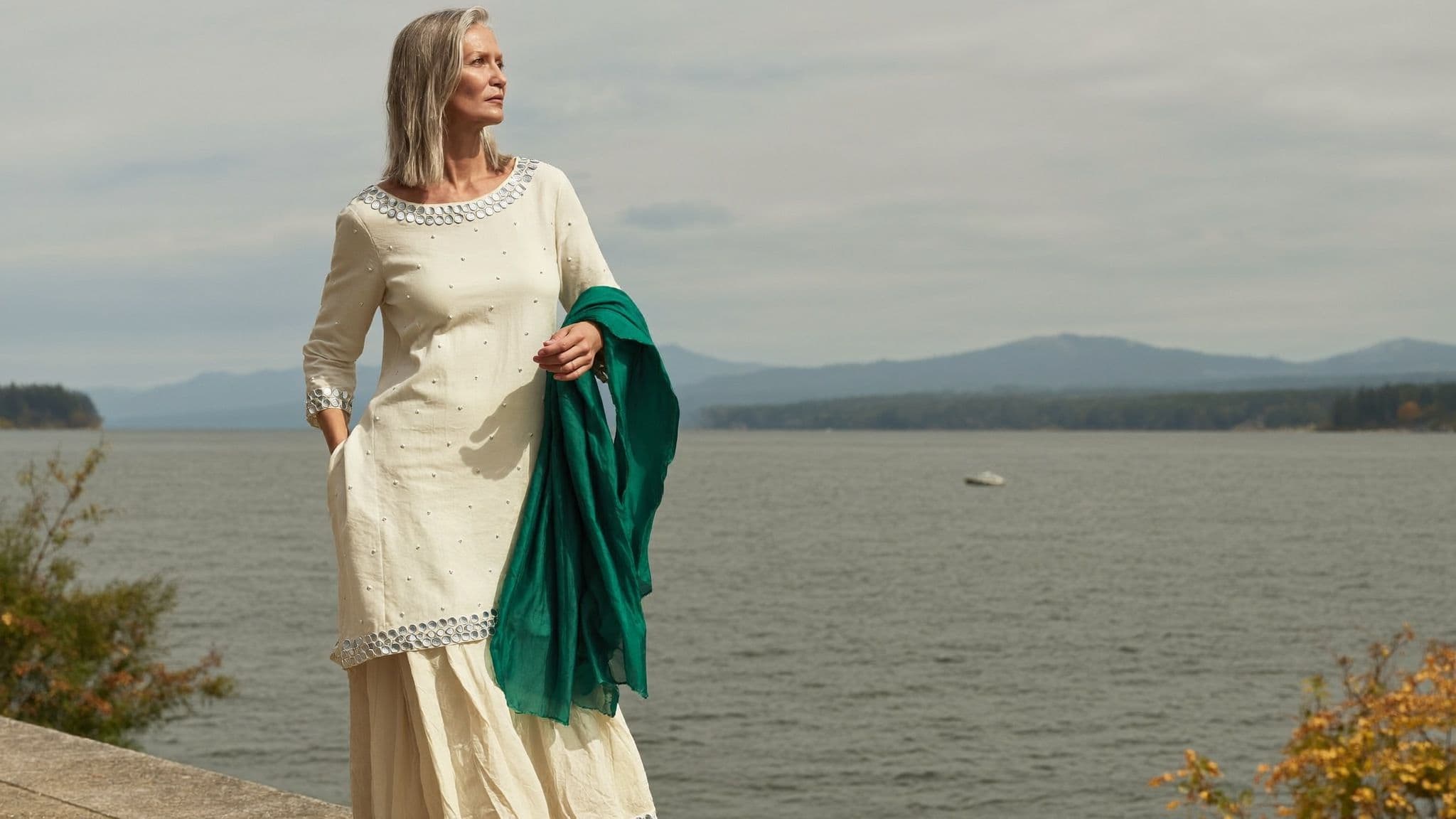 Woman in an ivory mirror-work kurta and sharara holding an emerald dupatta by the water