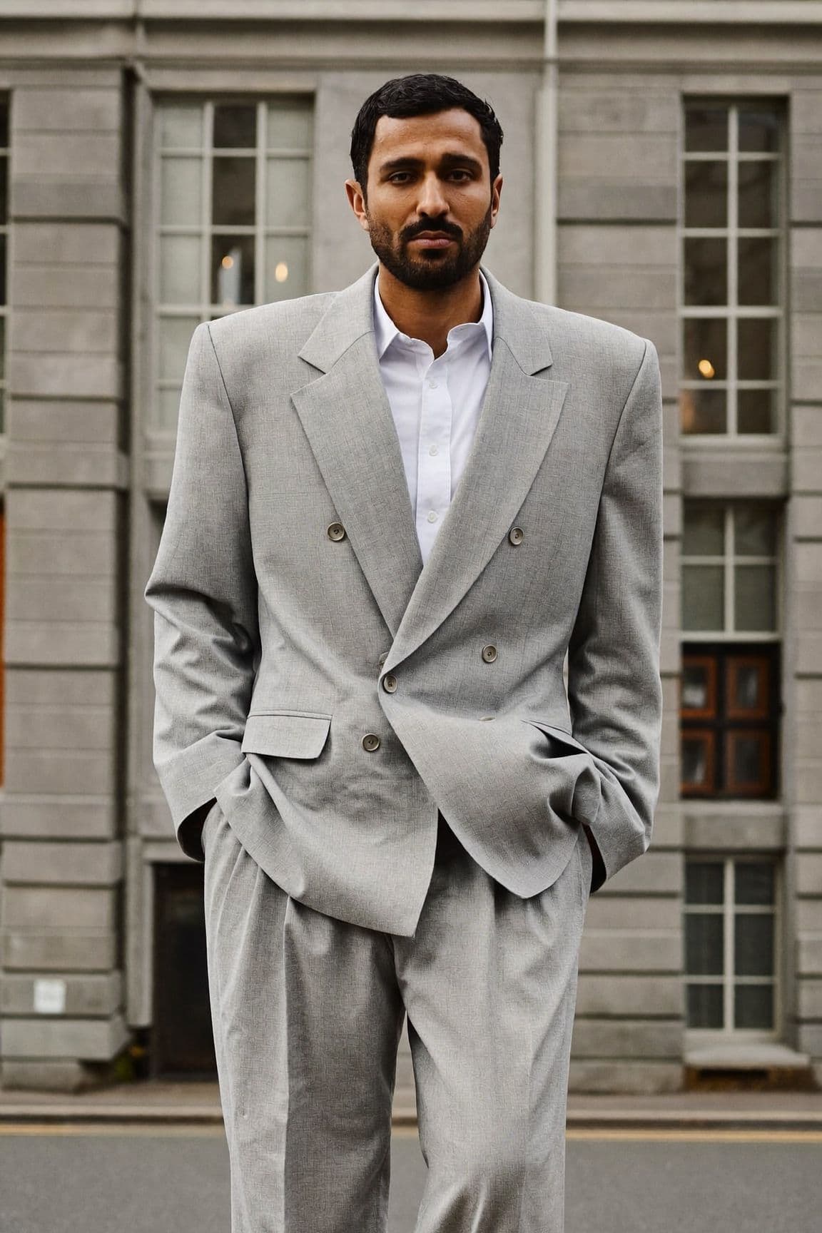 A man in an oversized light grey double-breasted suit with structured shoulders and wide pleated trousers, standing in front of a grey stone building