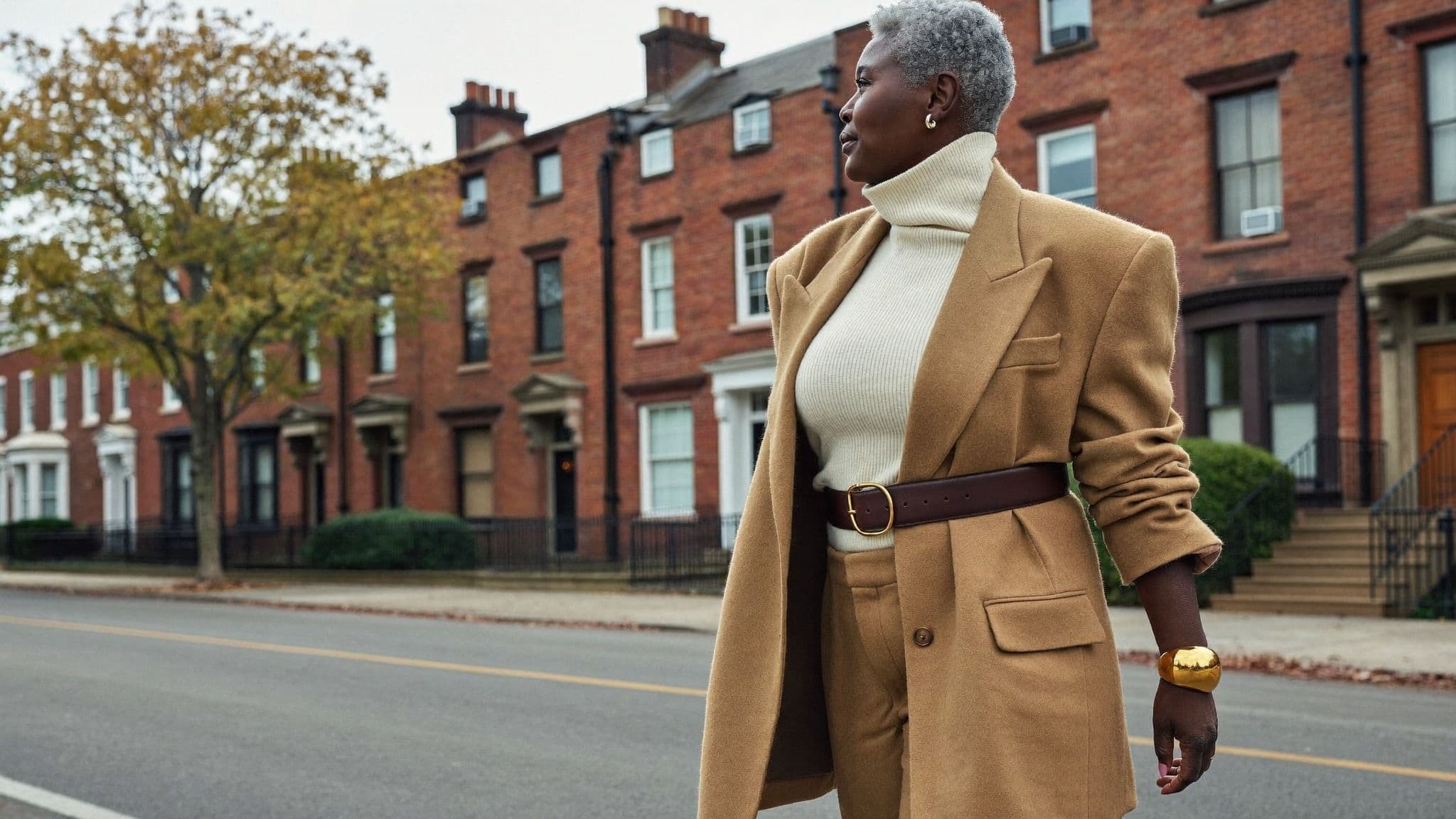 A woman with short grey hair walking past red-brick row houses in a camel blazer cinched with a wide brown leather belt, a cream funnel-neck knit, camel trousers and a gold cuff